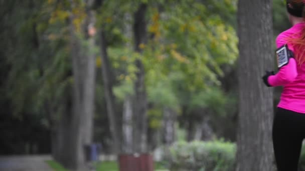 Runner giovane donna che corre nel parco autunnale esercitandosi all'aperto sotto la pioggia, tuta da allenamento rosa, vista posteriore, gadget sulla mano, rallentatore — Video Stock