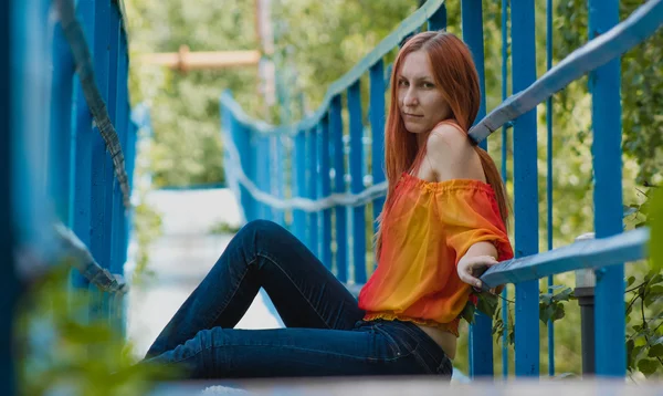 Young woman with red hair and yellow jacket sitting on a small bridge in summer day — Stock Fotó