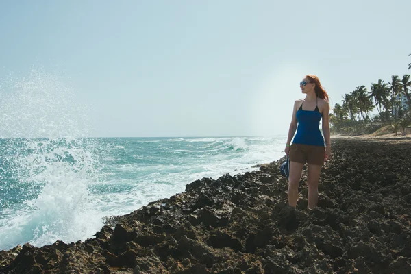 Jovem mulher em shorts e t-shirt em pé sobre rochas olhando para um mar — Fotografia de Stock