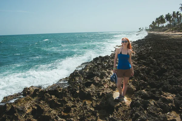 Young woman in in shorts and t-shirt standing on rocks  looking to a sea, the view from the top — Stock fotografie