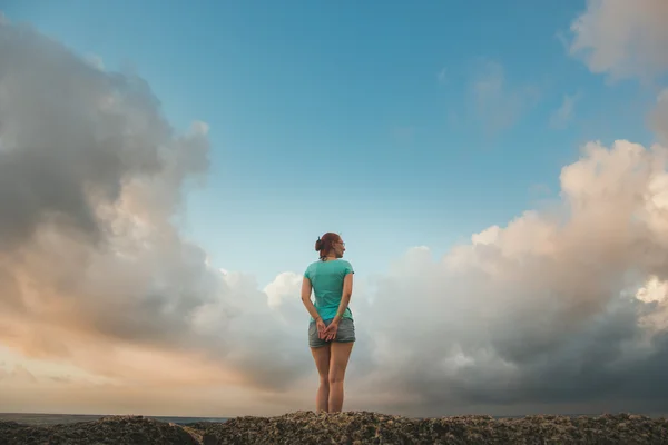 Молода жінка в шортах і синя футболка стоїть на узбережжі, дивлячись на море, хмари схожі на крила — стокове фото