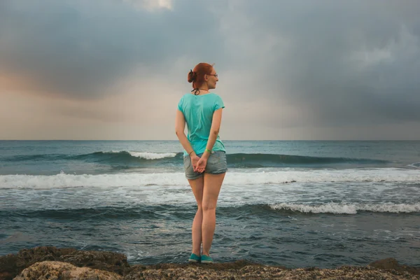 Jovem mulher em shorts e camiseta azul em pé na costa olhando para um mar — Fotografia de Stock