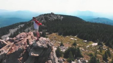 Elleri ayrık bir şekilde bir dağda duran genç adam manzaraya bakıyor.