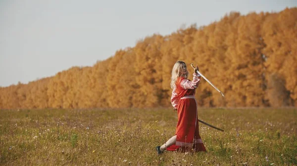Kırmızı elbiseli militan kadın sonbahar sahasında kılıç talimi yapıyor. — Stok fotoğraf