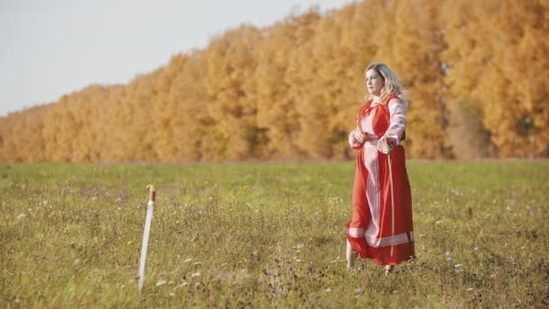 Training op het herfstveld - een strijdlustige vrouw in rode jurk hanteert een zwaard - nog een zwaard in de grond — Stockvideo