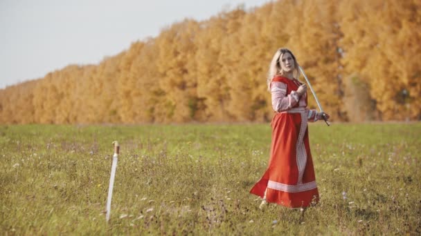 Treinamento no campo de outono - uma mulher beligerante de vestido nacional vermelho empunha com uma espada - outra espada presa no chão — Vídeo de Stock