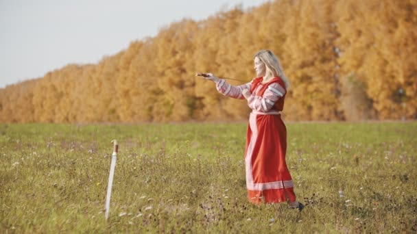 Training op het herfstveld - een strijdlustige blonde vrouw in rode nationale kleding hanteert een zwaard - een ander zwaard in de grond — Stockvideo