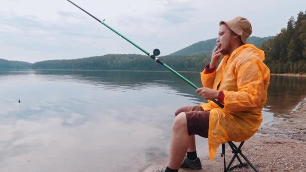 Uomo solitario in impermeabile arancione seduto su una riva e fumare una sigaretta sulla pesca — Video Stock