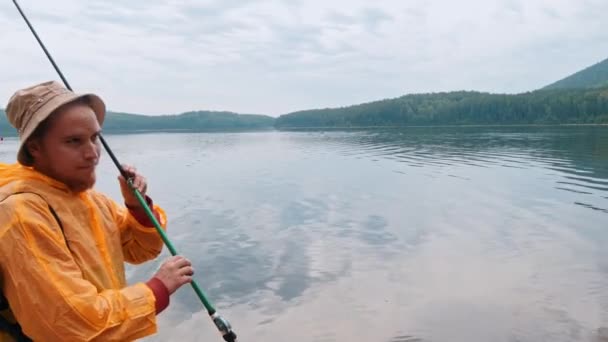 Pescador sonriente en un impermeable de pie en una orilla sosteniendo su caña de pescar — Vídeos de Stock