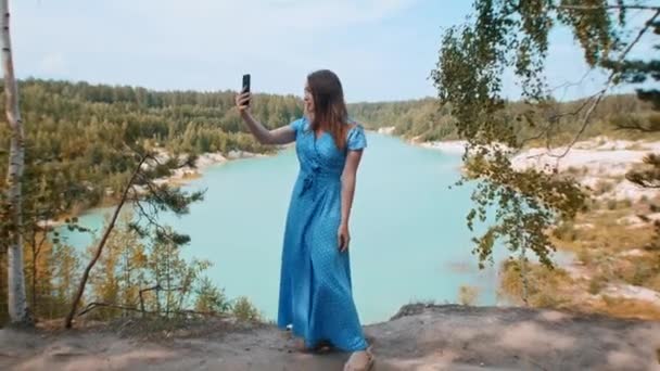 Junge Frau in blauem Kleid steht vor einem Hintergrund aus blauem See und Wald und macht ein Selfie — Stockvideo