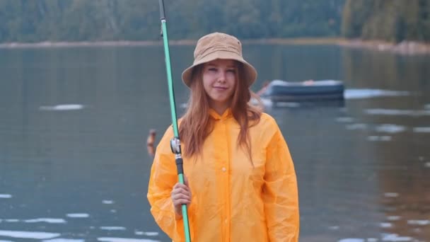 Mladá žena v oranžovém plášti a klobouku na rybaření - dívá se do kamery — Stock video
