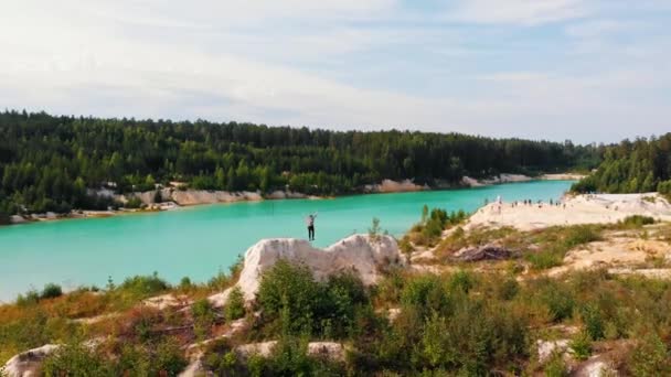 Landschaft eines hellblauen schlammigen Sees umgeben von Wald - ein erfolgreicher Mann läuft auf einen Felsen und hebt die Hände — Stockvideo