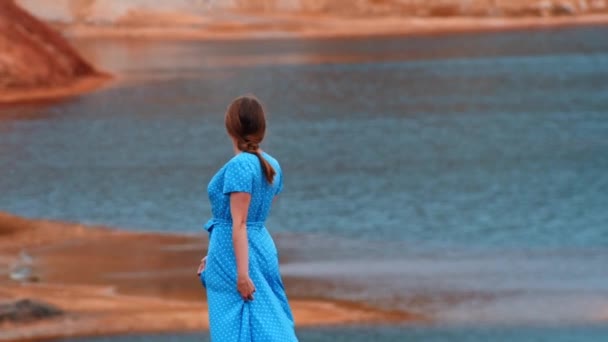 Jonge vrouw in blauwe jurk dansen op een achtergrond van een oranje klei strand en meer — Stockvideo