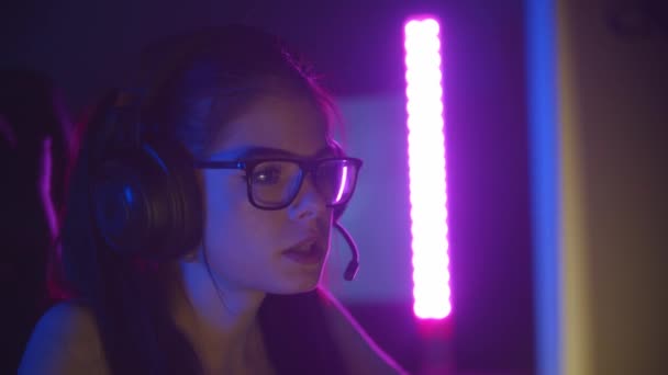 Mujer bonita joven en gafas jugando juegos en línea en el club de juegos de neón y hablando en el micrófono — Vídeo de stock