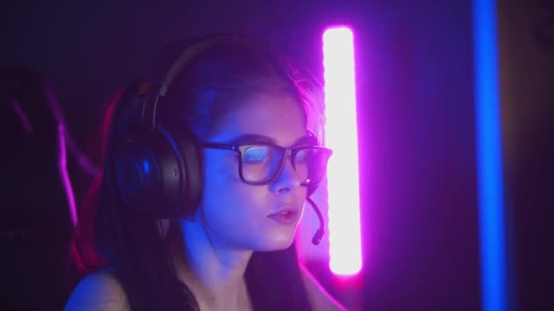 Mujer bonita joven en gafas jugando juegos en línea en el club de juegos de neón - usando auriculares y hablando en el micrófono — Vídeo de stock