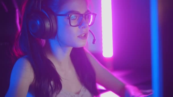 Fiatal nő szemüvegben játszik online játékok neon játék klub - visel fejhallgató és beszél a mikrofonban — Stock videók