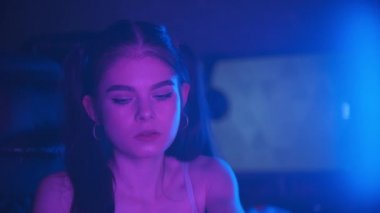Genç çekici bir kadın neon oyun kulübünde oturuyor ve büyük kulaklıklar takıyor ve takımını çevrimiçi oyunda selamlıyor.