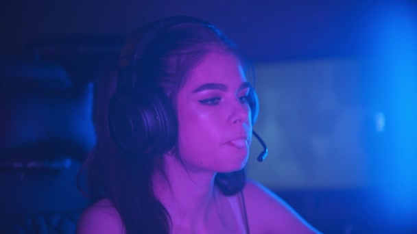 Junge hübsche Frau sitzt in einem neonfarbenen Spielclub und bläst während der Spielsitzung einen Blasenkaugummi — Stockvideo