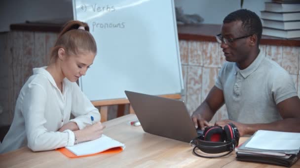 Lección de inglés - una mujer y un hombre afroamericano teniendo una clase de inglés - un hombre sentado junto a la computadora portátil y una mujer tomando notas — Vídeos de Stock