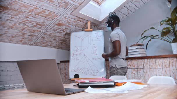 Une leçon de mathématiques en ligne - un professeur afro-américain en masque de médecine debout près du tableau et explique le matériel pour ses élèves devant l'ordinateur portable — Video