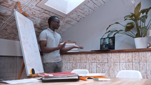 Une leçon de mathématiques en ligne - un enseignant afro-américain dans des lunettes debout près du tableau et explique le matériel pour ses élèves sur l'écran d'un ordinateur portable — Video