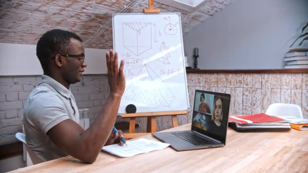 Une leçon de mathématiques en ligne - un enseignant afro-américain en lunettes saluant ses élèves à l "écran — Video