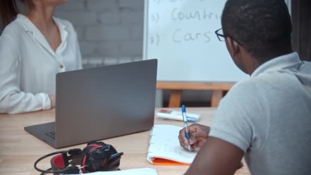 Profesora dándole a un hombre negro una lección de inglés - apuntando al tablero con temas para la primera lección — Vídeos de Stock