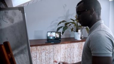 Çevrimiçi bir ders. Gözlüklü Afro-Amerikan bir erkek öğretmen tahtada duruyor ve öğrencilerine ekrandaki materyalleri anlatıyor.
