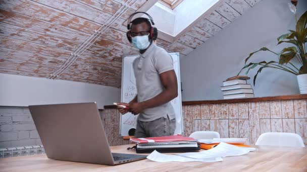 An online maths lesson - an african-american man teacher in medicine mask and big headphones standing by the board and explains the material for his students in front of the laptop — Stock Video