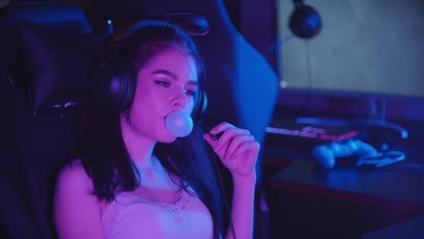 Genç bir kadın neon oyun kulübünde oturuyor ve sakız üflüyor. Büyük kulaklıklar takıyor ve mikrofonla konuşuyor. — Stok video