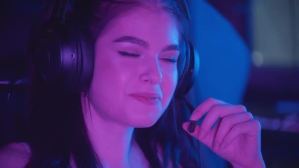 Jovem mulher bonita sentada no clube de jogos de néon vestindo grandes fones de ouvido e falando em microfone — Vídeo de Stock