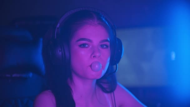 Junge hübsche Frau sitzt in einem neonfarbenen Spielclub und pustet einen Blasengummi - schaut in die Kamera — Stockvideo
