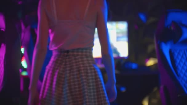 Fiatal csinos nő besétál a neon játék klub és leül a székre - felveszi a fejhallgatót, és elkezd játszani a játékot — Stock videók