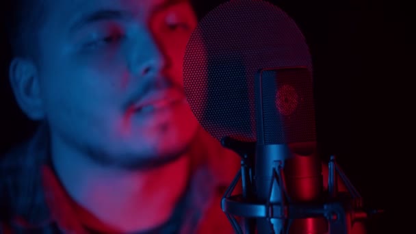 ミュージシャンのコンセプト-男は赤と青の照明で暗いスタジオに座って、ポップフィルターでマイクで歌を歌います — ストック動画