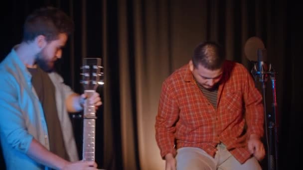 Dois homens sentam-se em cadeiras no estúdio de gravação e gravam uma música - um deles pega uma guitarra — Vídeo de Stock