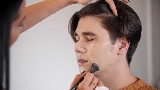 女性は、ブラシを使用して男性モデルの顔に厚い基盤を適用するアーティストを構成します。 — ストック動画