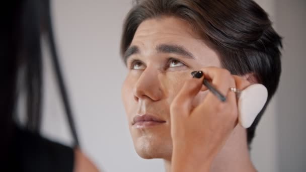 Mujer maquillaje artista aplicando corrector bajo los ojos de modelo masculino usando un cepillo — Vídeos de Stock
