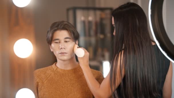 Woman make up artist applying foundation on the face of male model using a wet sponge - mirror reflection — Stock Video