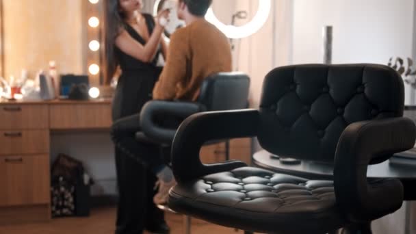 Maquiagem masculina modelo masculino sentado na cadeira e artista mulher aplicando maquiagem em seu rosto — Vídeo de Stock
