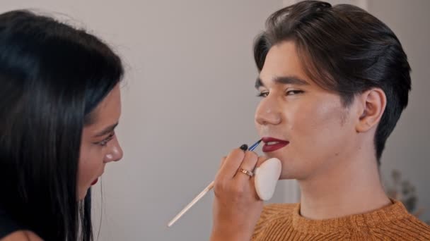 Makijaż studio - makijaż artysty stosującego czerwoną matową szminkę na ołówku na ustach męskiego modelu za pomocą pędzla — Wideo stockowe