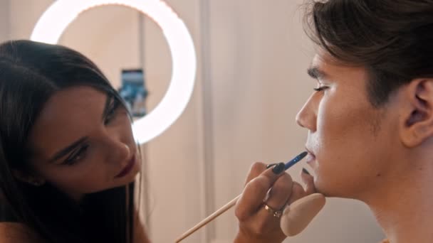 Estudio de maquillaje - artista de maquillaje que aplica lápiz labial rojo sobre el lápiz en los labios de un guapo modelo masculino usando un pincel — Vídeos de Stock