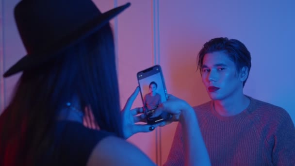 Makeup studio - γυναίκα καλλιτέχνης λήψη φωτογραφιών του ανδρικού μοντέλου της με μακιγιάζ σε νέον φωτισμό - τολμηρή εμφάνιση με κόκκινα χείλη — Αρχείο Βίντεο
