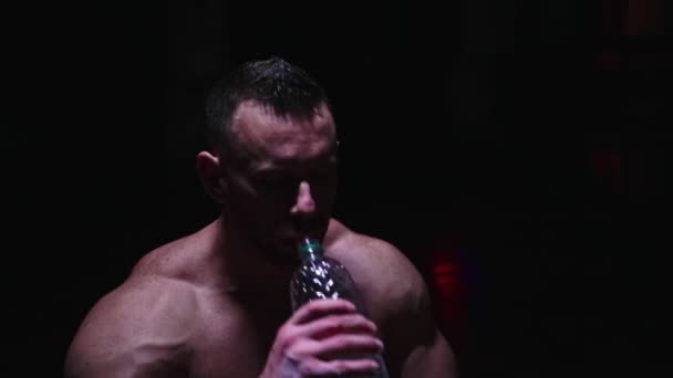 Сильный мужчина без рубашки пьет воду из бутылки в темноте — стоковое видео