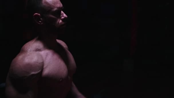 Fuerte hombre sin camisa bombeando sus bíceps en el cuarto oscuro — Vídeos de Stock