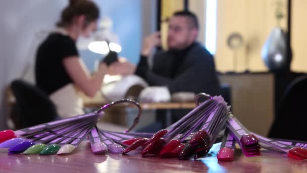 Manicure salon - nail polish samples on the foreground - a man having his nails done on the background — Stock Video