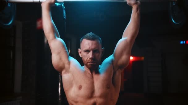 Hombre duro de pie en el gimnasio y levanta una barra pesada y bombea los músculos de sus manos sobre su cabeza — Vídeos de Stock