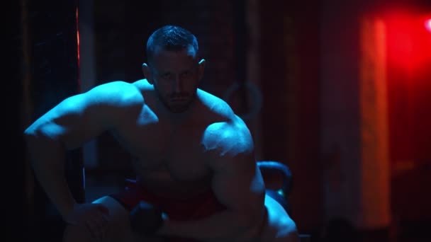 Homem forte bombeando seus bíceps em iluminação de contraste e olhando para a câmera — Vídeo de Stock