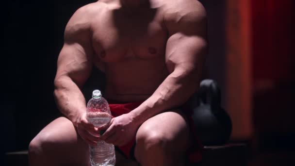 Homem duro sentado no ginásio e segurando garrafa de água de plástico — Vídeo de Stock