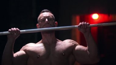 Spor salonunda terli, sert, üstsüz bir adam ağır bir çubuğu çekip bağırıyor.