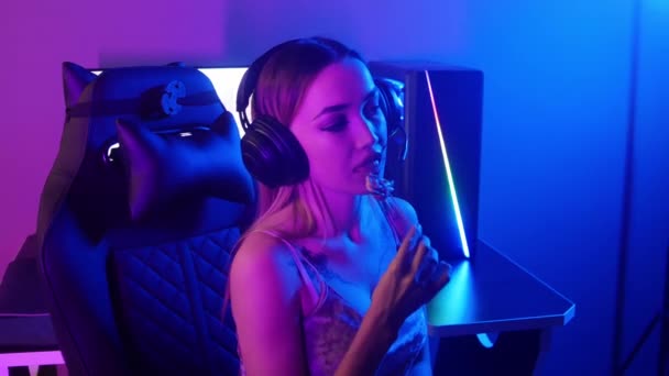 Young blonde gamer woman sits in the chair in neon gaming club and sucking a lollipop — Stock Video